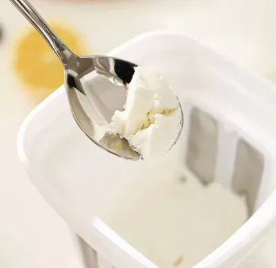 A metal spoon scooping thick cream cheese or strained yogurt out of a white Greek Yogurt Strainer basket.