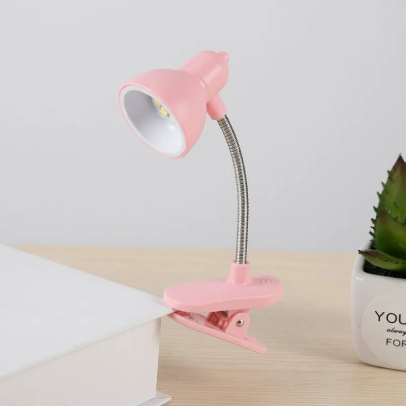 Pink Mini Clip Study Lamp attached to a book cover on a wooden desk with flexible silver neck bent.